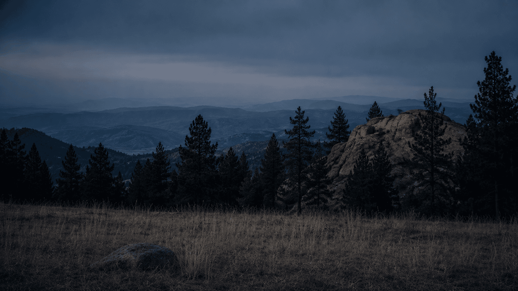Colorado high foothills at dusk — pines, granite, layered ridges receding into atmospheric haze.